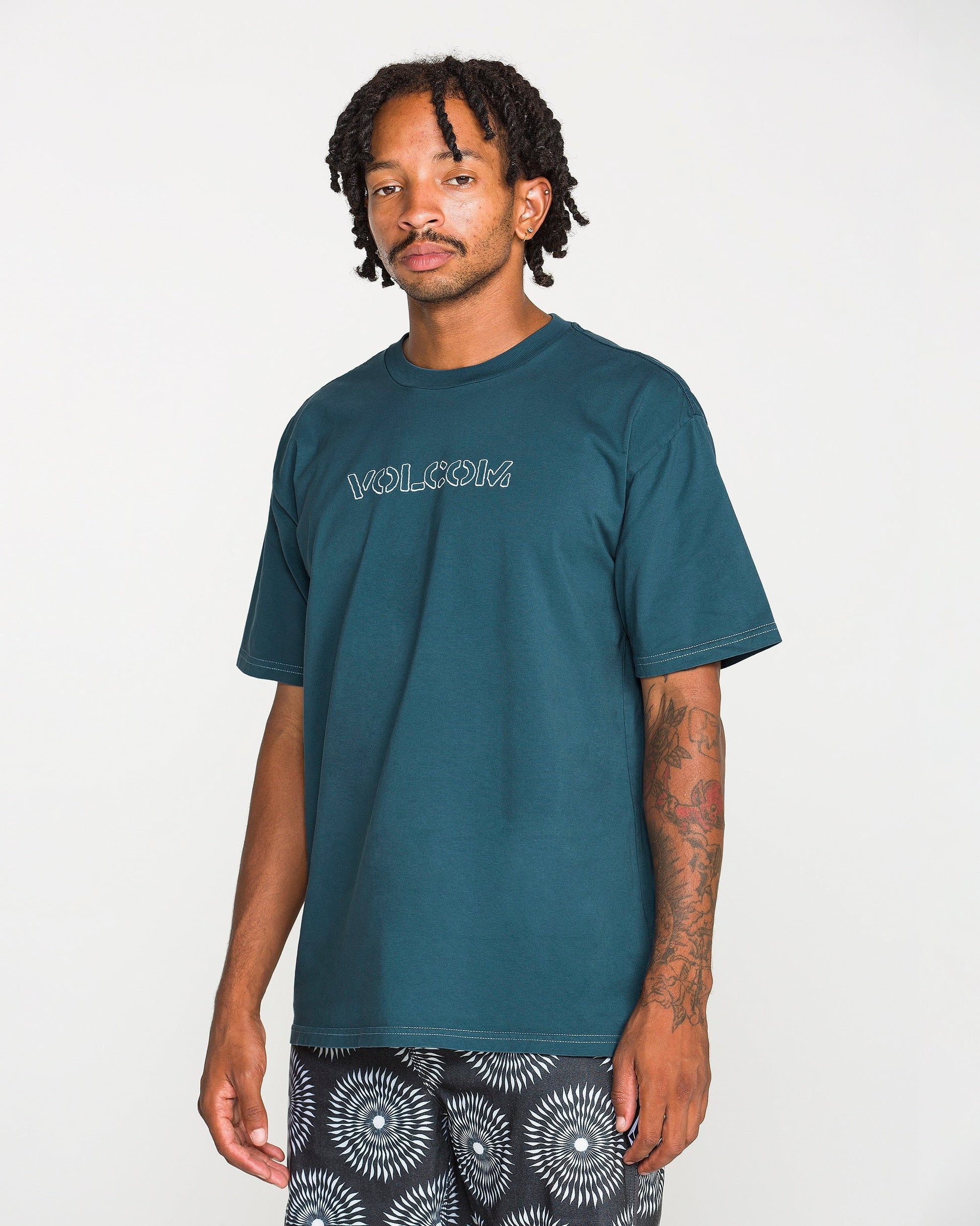 A man with short dreadlocks and arm tattoos wears the Volcom Men's Crassy Short Sleeve Tee in teal, featuring a soft-hand screenprint, paired with black-and-white patterned shorts and standing against a plain white background.