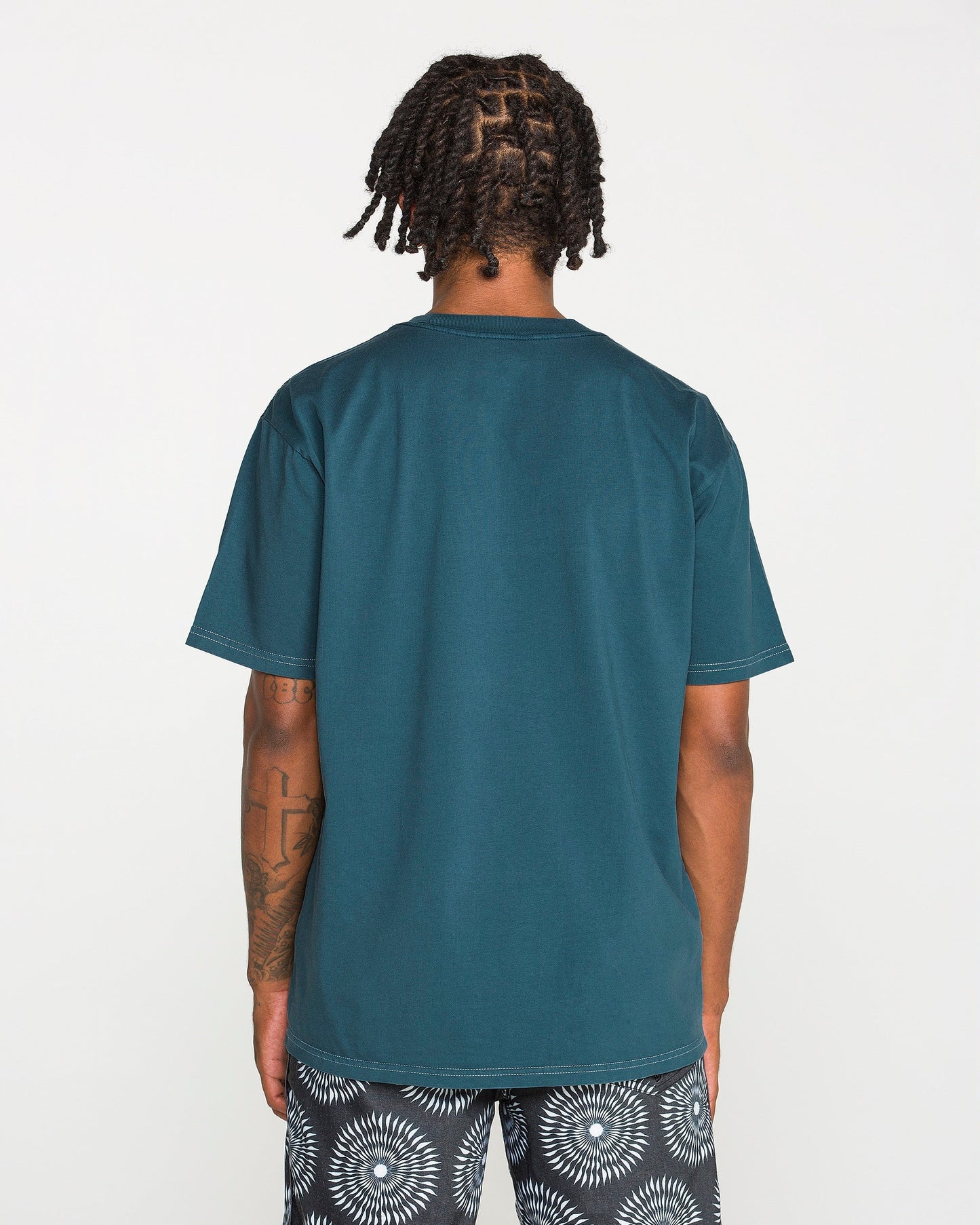 A person with short dreadlocks is standing with their back to the camera, wearing the Volcom Men's Crassy Short Sleeve Tee in teal paired with patterned black-and-white shorts. The loose fit of the t-shirt drapes comfortably, set against a plain white background.