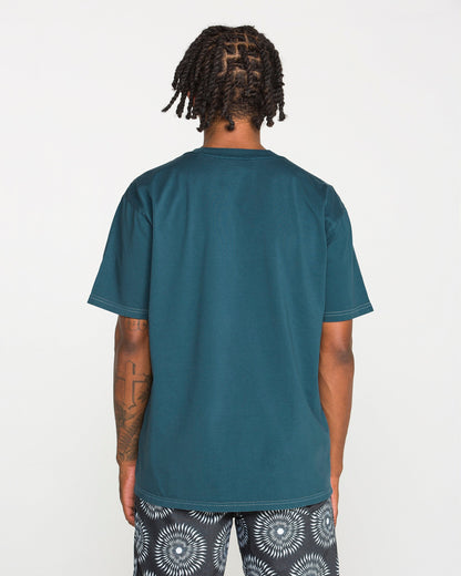 A person with short dreadlocks is standing with their back to the camera, wearing the Volcom Men's Crassy Short Sleeve Tee in teal paired with patterned black-and-white shorts. The loose fit of the t-shirt drapes comfortably, set against a plain white background.
