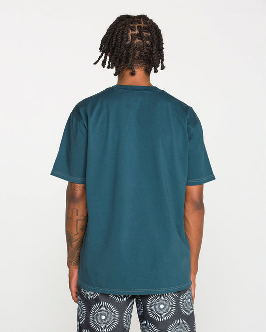 A person with short dreadlocks is standing with their back to the camera, wearing the Volcom Men's Crassy Short Sleeve Tee in teal paired with patterned black-and-white shorts. The loose fit of the t-shirt drapes comfortably, set against a plain white background.