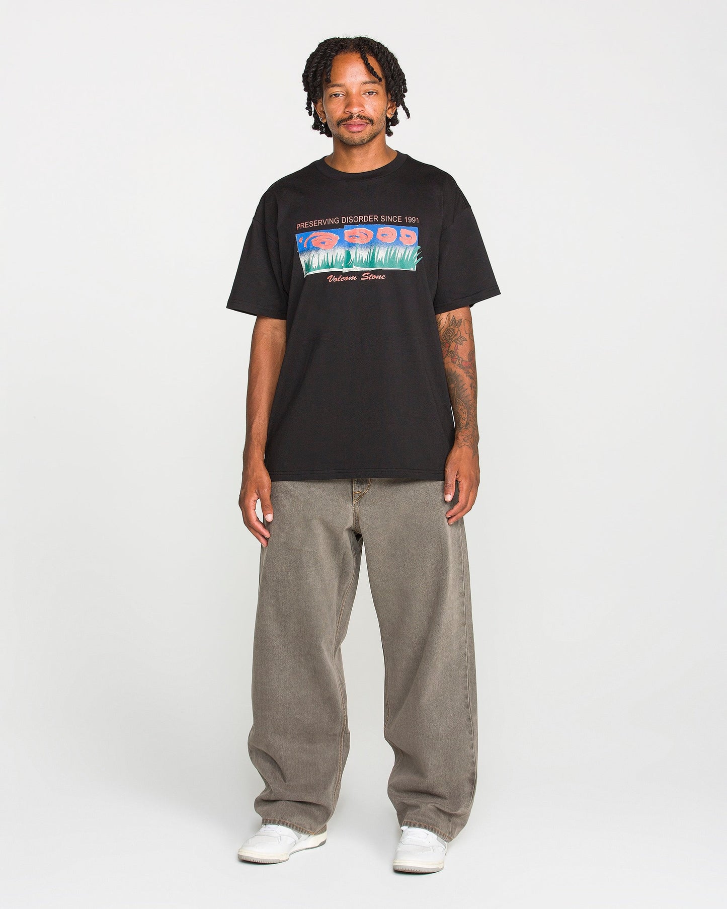 A man with short dreadlocks wears the Volcom Men's Featured Artist Antonio Carrau Short Sleeve Tee—a premium black t-shirt showcasing a bold graphic design—paired with baggy gray jeans and white sneakers, standing facing forward against a plain white background.