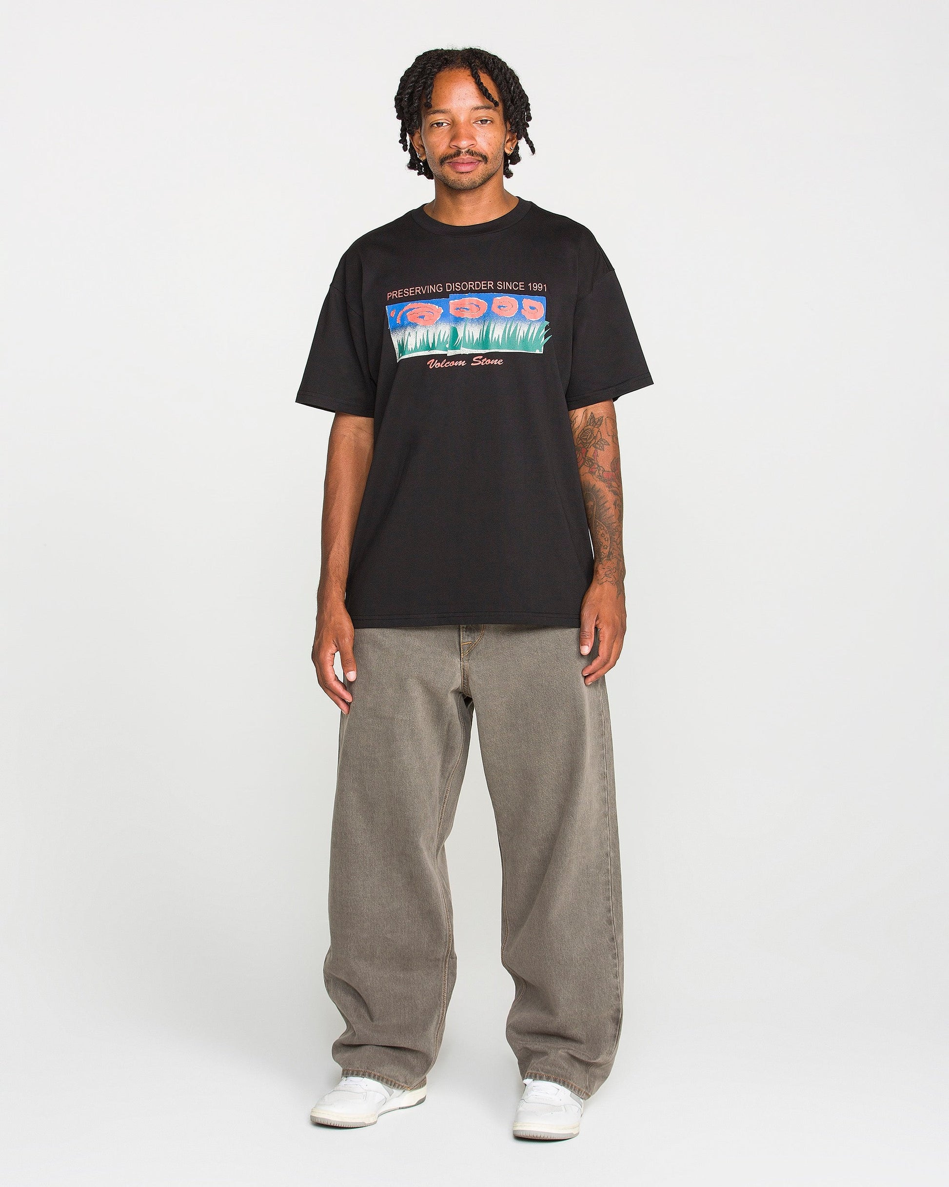 A man with short dreadlocks wears the Volcom Men's Featured Artist Antonio Carrau Short Sleeve Tee—a premium black t-shirt showcasing a bold graphic design—paired with baggy gray jeans and white sneakers, standing facing forward against a plain white background.