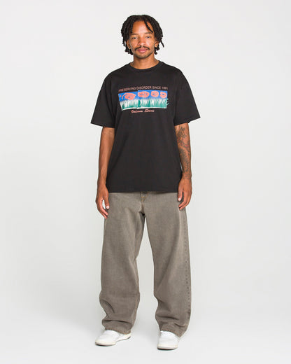 A man with short dreadlocks wears the Volcom Men's Featured Artist Antonio Carrau Short Sleeve Tee—a premium black t-shirt showcasing a bold graphic design—paired with baggy gray jeans and white sneakers, standing facing forward against a plain white background.