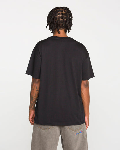 A person with short dreadlocks is standing with their back to the camera against a plain white background, wearing the Volcom Men's Featured Artist Antonio Carrau Short Sleeve Tee in black and gray jeans.