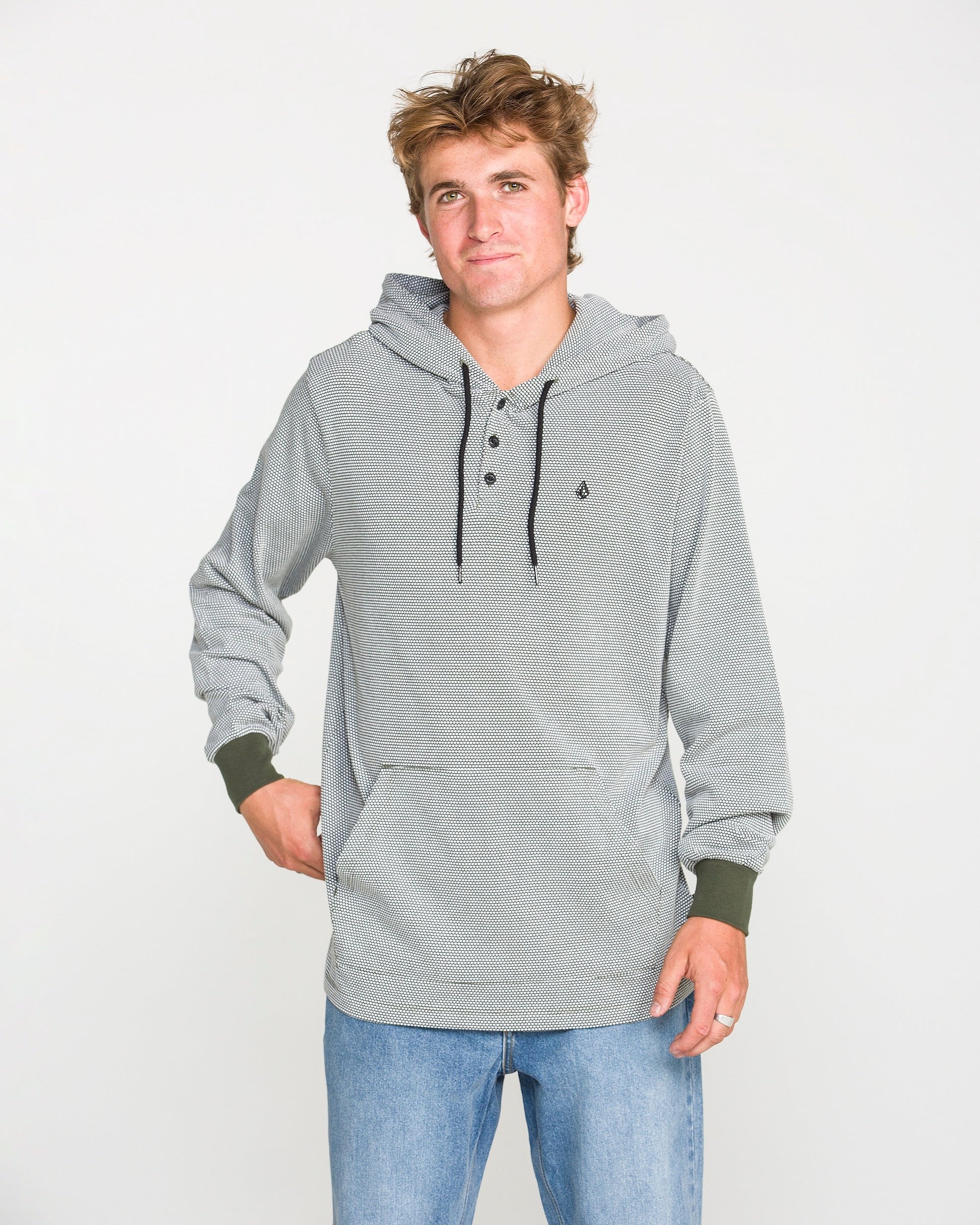 A young man wearing the Volcom Men's Rowan Thermal Long Sleeve Shirt in light gray, featuring black drawstrings, pairs it with blue jeans as he stands against a plain white background, looking at the camera with a relaxed expression.