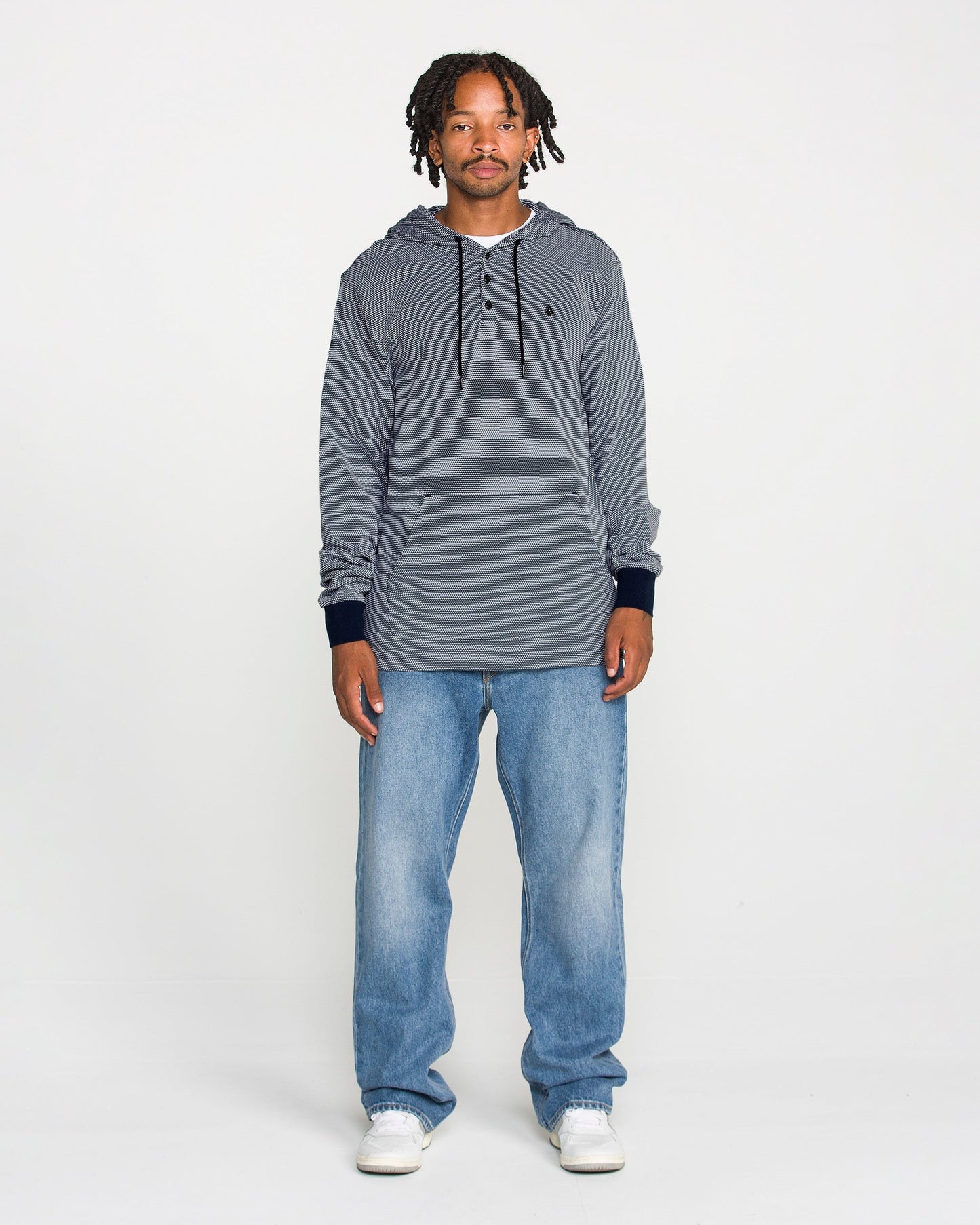 A man with short dreadlocks stands against a plain white background, wearing the Volcom Men's Rowan Thermal Long Sleeve Shirt for midweight warmth, paired with light blue jeans and white sneakers. He looks directly at the camera with a neutral expression.