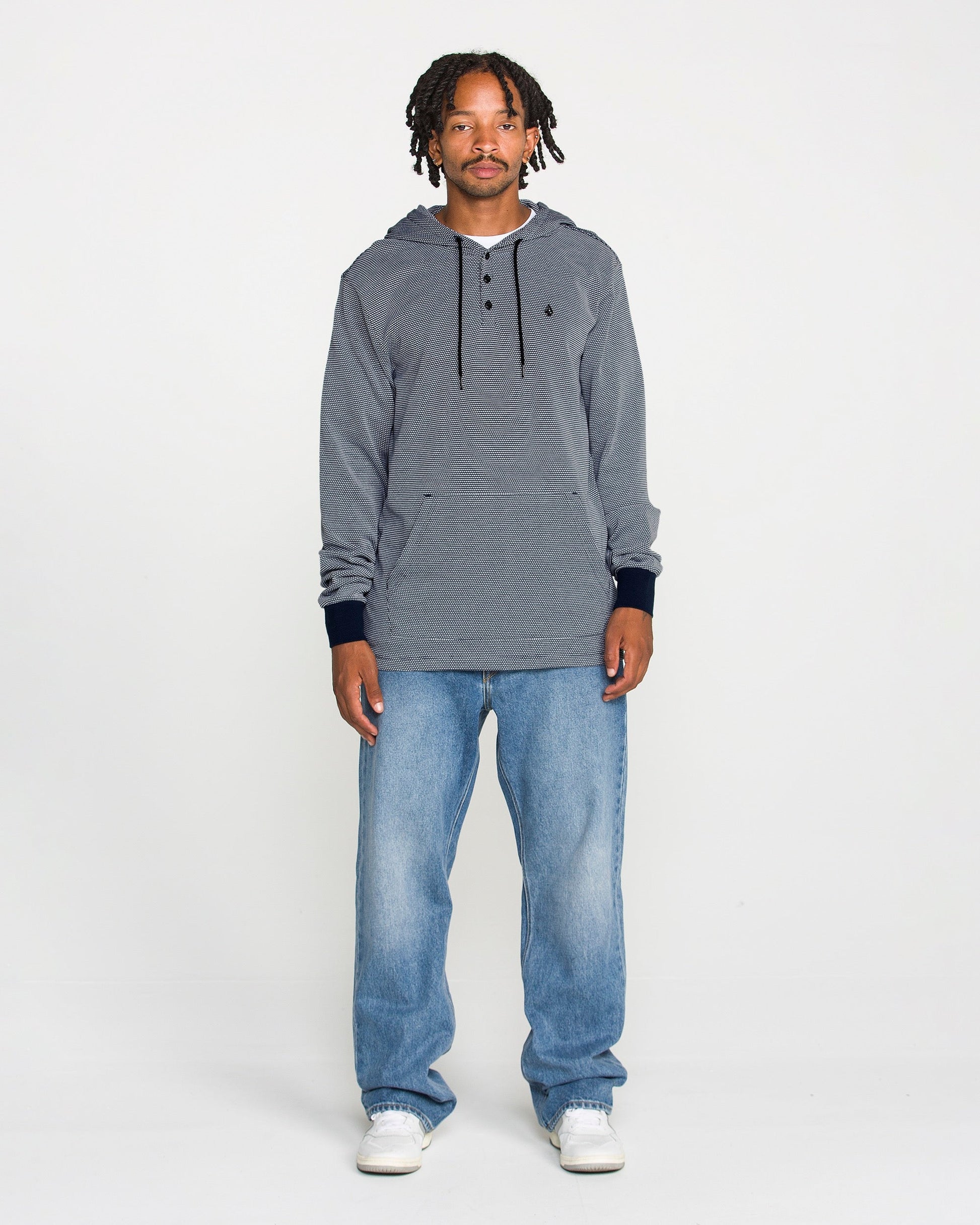A man with short dreadlocks stands against a plain white background, wearing the Volcom Men's Rowan Thermal Long Sleeve Shirt for midweight warmth, paired with light blue jeans and white sneakers. He looks directly at the camera with a neutral expression.