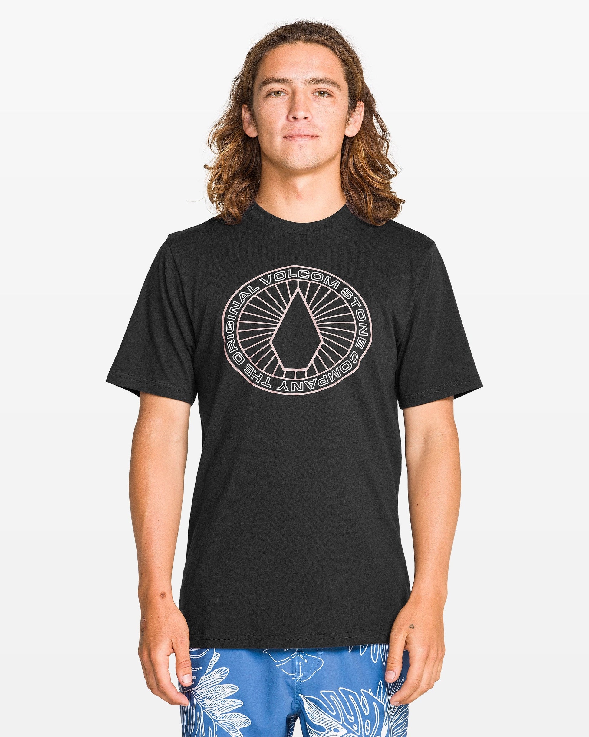A person with long brown hair wears a Volcom Men's Medalist Short Sleeve Tee with a modern fit and circular geometric graphic, styled with blue shorts featuring a white leaf print. The ring-spun heather fabric adds comfort against a plain background.