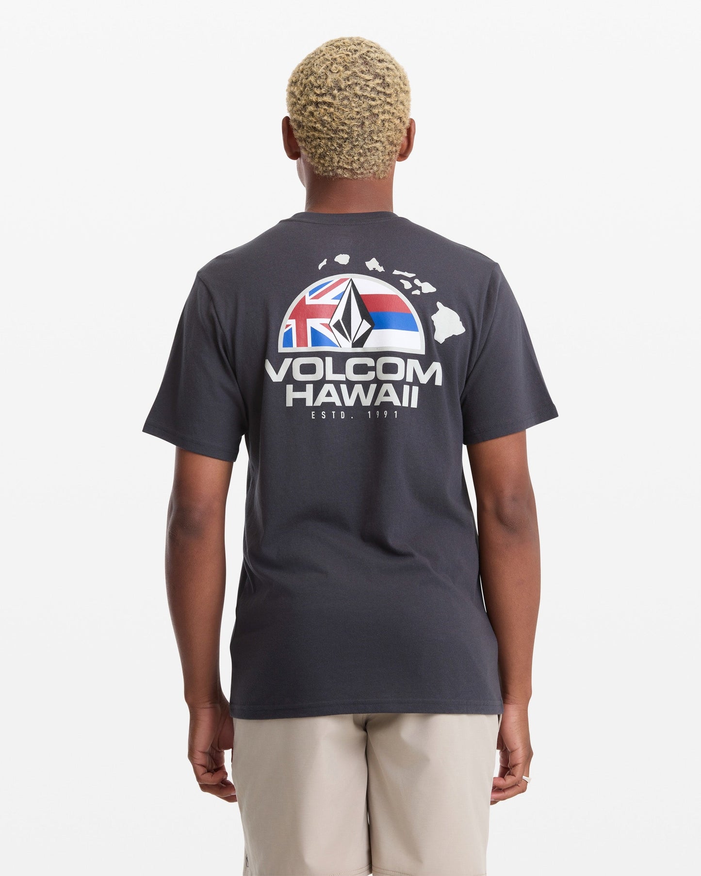 A man with short blond hair wears the Volcom Men's Allegiance Short Sleeve Tee, featuring "VOLCOM HAWAII" text, a Hawaii outline, and flag graphic on the back, standing against a plain white background.