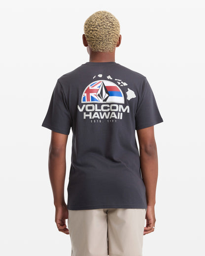 A man with short blond hair wears the Volcom Men's Allegiance Short Sleeve Tee, featuring "VOLCOM HAWAII" text, a Hawaii outline, and flag graphic on the back, standing against a plain white background.