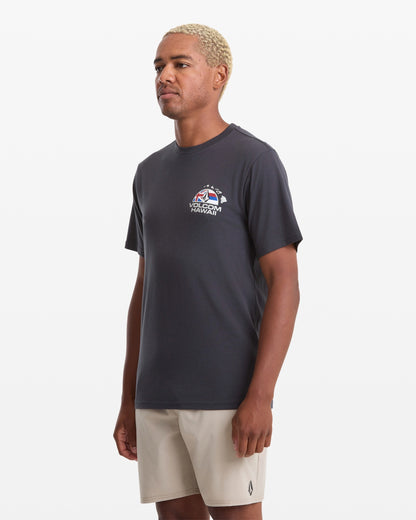 A man with short blond hair wears a dark gray Volcom Men's Allegiance Short Sleeve Tee, featuring a small chest graphic, and light beige shorts while standing against a plain white background.