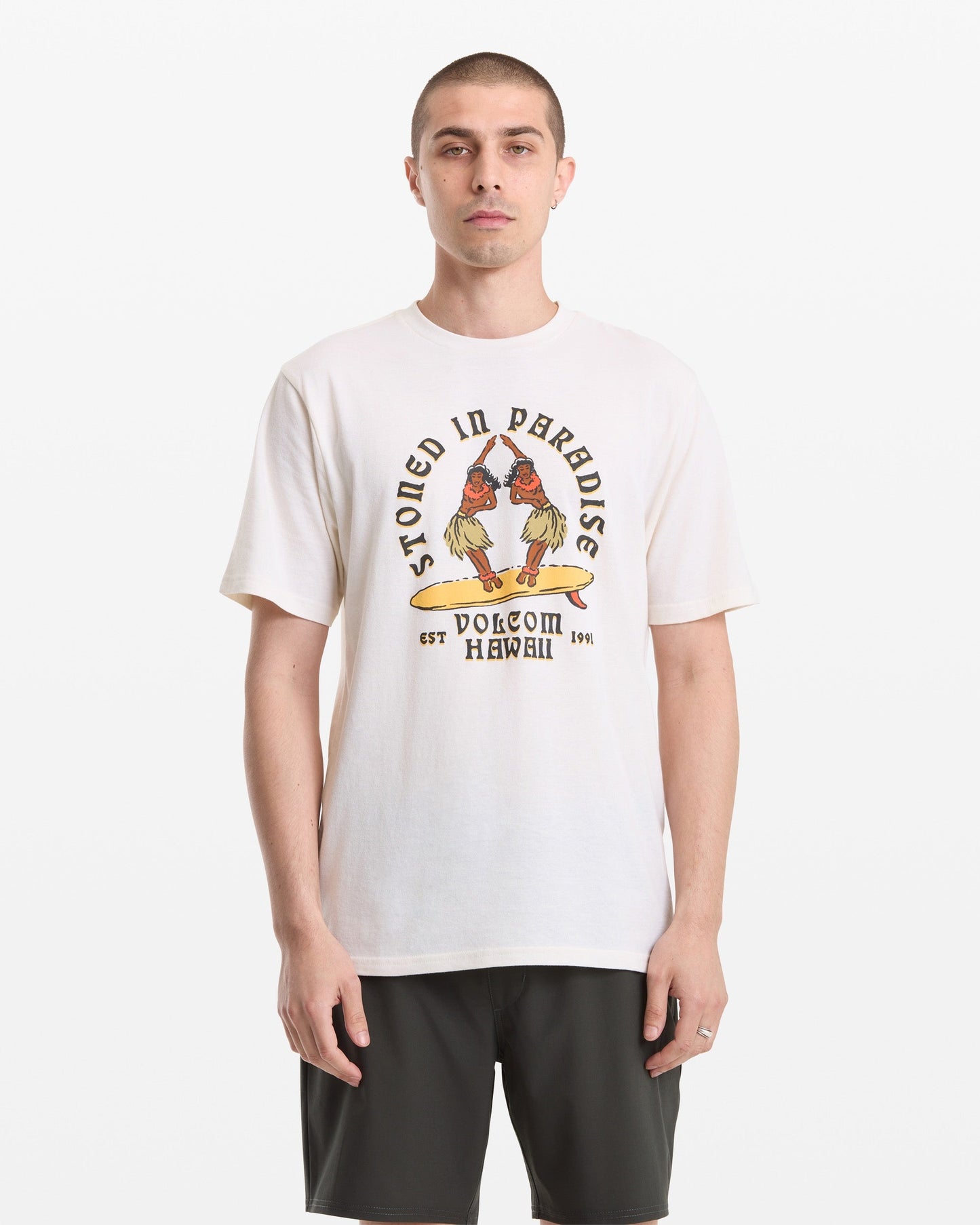 A man with short hair wears the Volcom Men's Stoned In Paradise Short Sleeve Tee, featuring "Stoned in Paradise" text and parrot graphic, paired with dark shorts, standing against a plain white background.