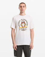 A man with short hair wears the Volcom Men's Stoned In Paradise Short Sleeve Tee, featuring "Stoned in Paradise" text and parrot graphic, paired with dark shorts, standing against a plain white background.