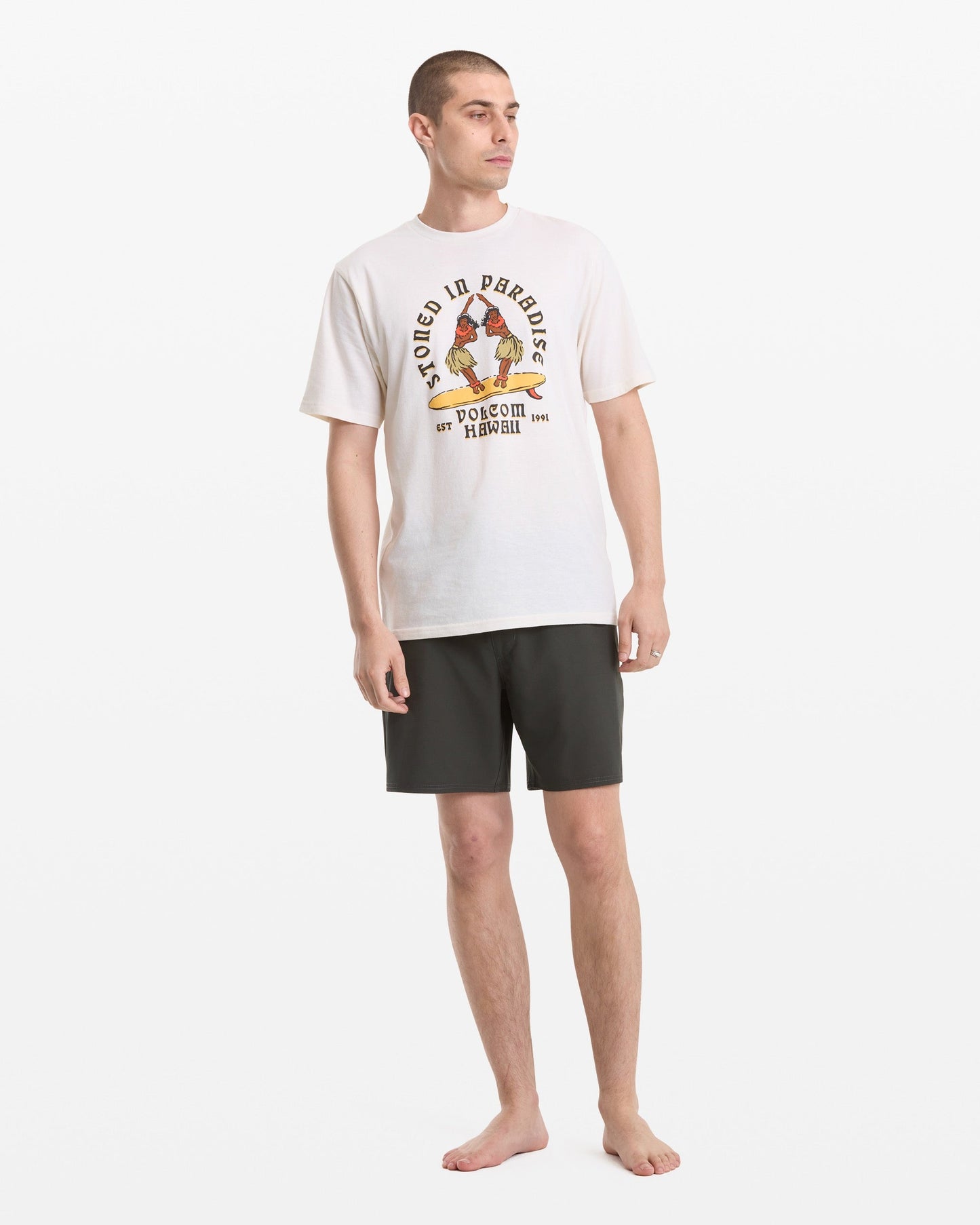 A barefoot man stands against a white background, wearing the Volcom Men's Stoned In Paradise Short Sleeve Tee with a colorful graphic and black shorts. He looks slightly to the side, arms relaxed by his sides.