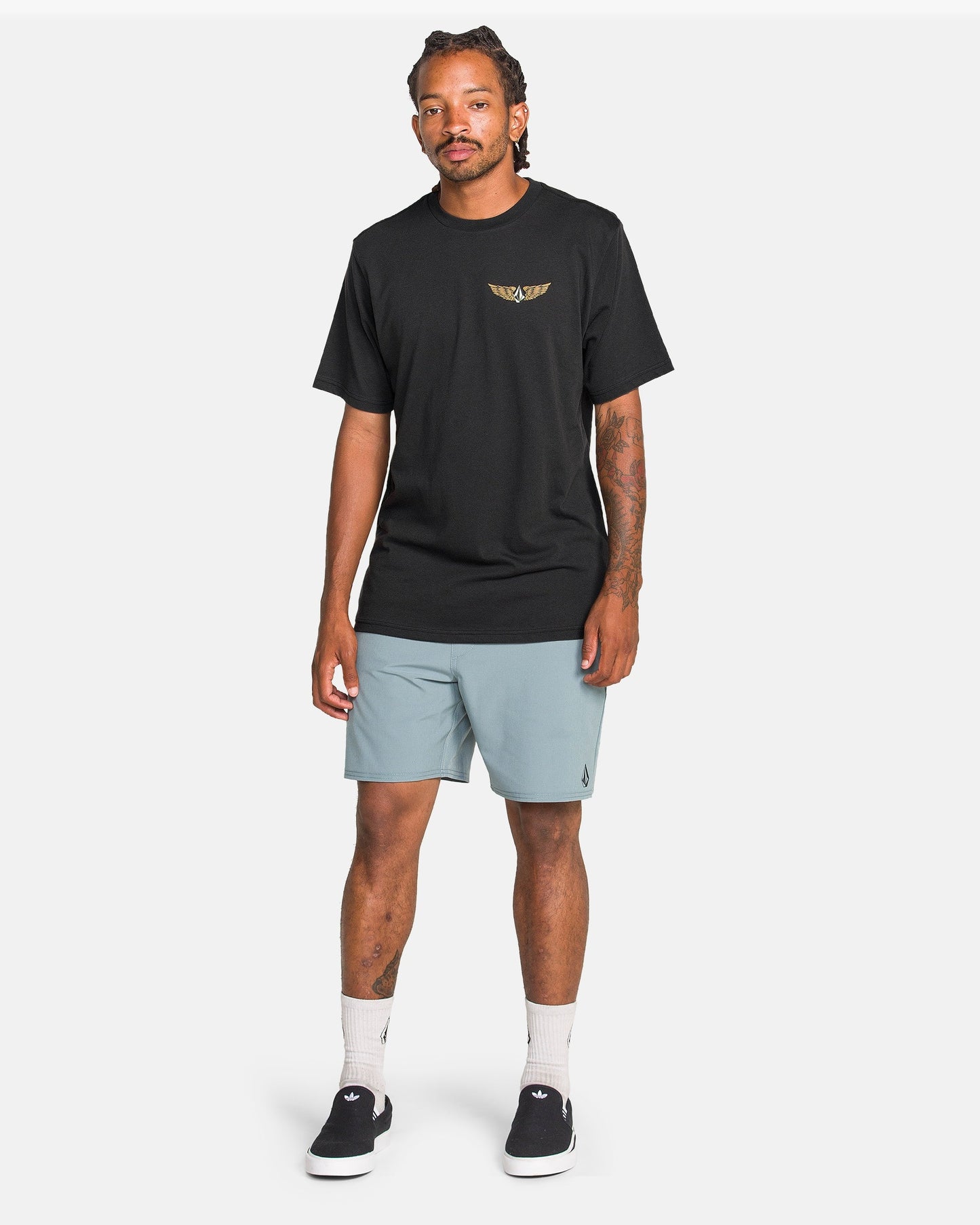 A man with styled hair and forearm tattoos stands against a white background wearing a black Volcom Men's Hi Flyer Short Sleeve Tee, light blue shorts, white socks, and black slip-on shoes.