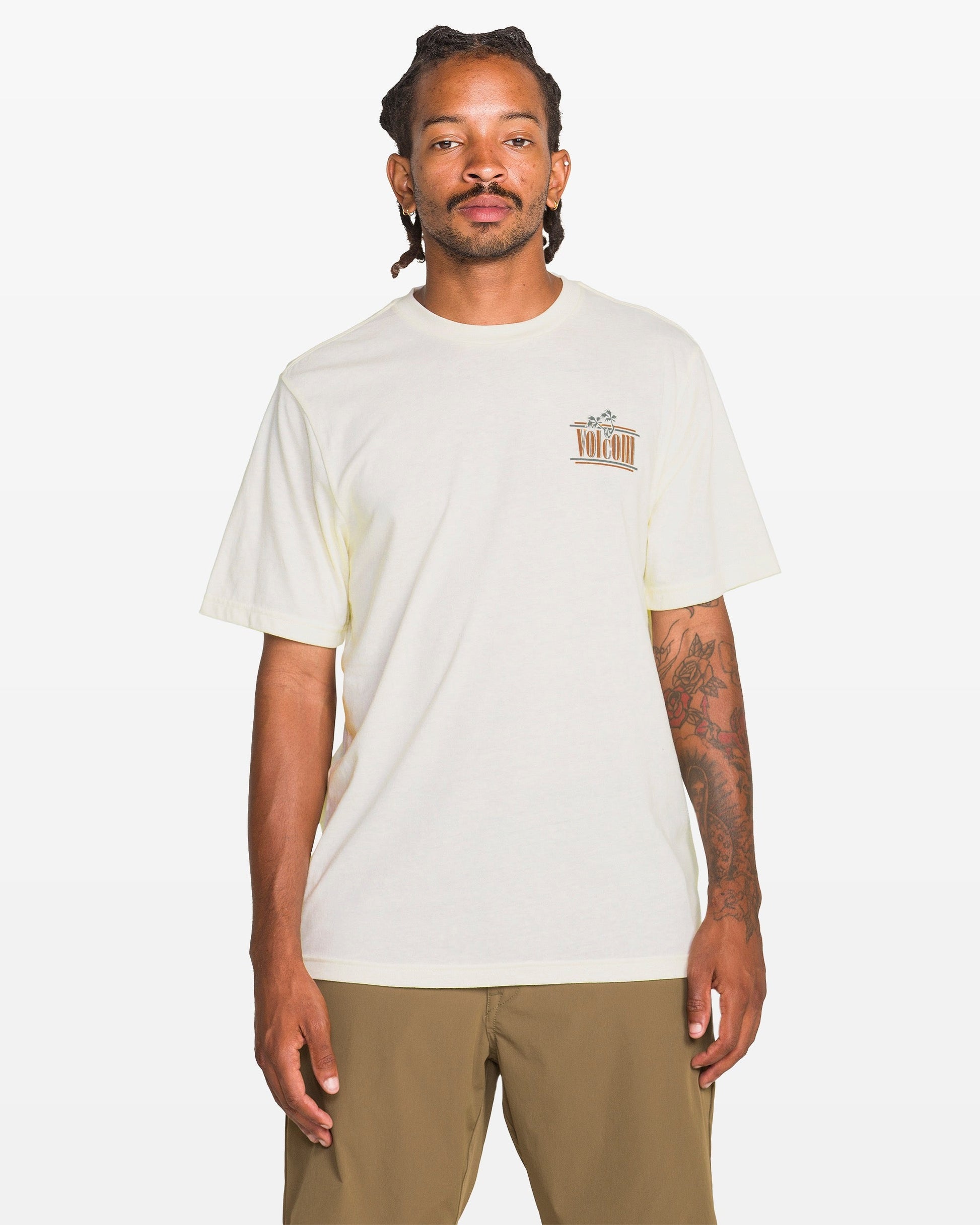 A person with dreadlocks and tattoos on their right arm wears the Volcom Men's Breathe Deep Short Sleeve Tee in heather, featuring a small left chest graphic, paired with khaki shorts, standing against a plain white background.