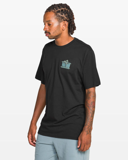 A man with braided hair and arm tattoos wears a Volcom Men's Breathe Deep Short Sleeve Tee featuring a small blue chest graphic and light blue shorts, standing against a plain white background.