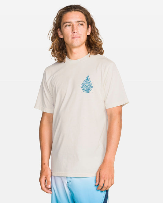 A person with long brown hair is wearing the Volcom Men's All Seeing Short Sleeve Tee in Off White Heather, which has a blue geometric droplet design, and blue gradient shorts while standing against a plain white background.