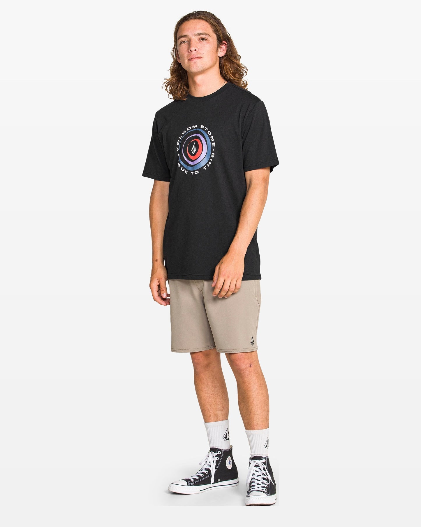 A young man with long hair faces forward, wearing the Volcom Men's Enterprise Short Sleeve Tee in black, paired with beige shorts, white socks, and black high-top sneakers against a plain white background.