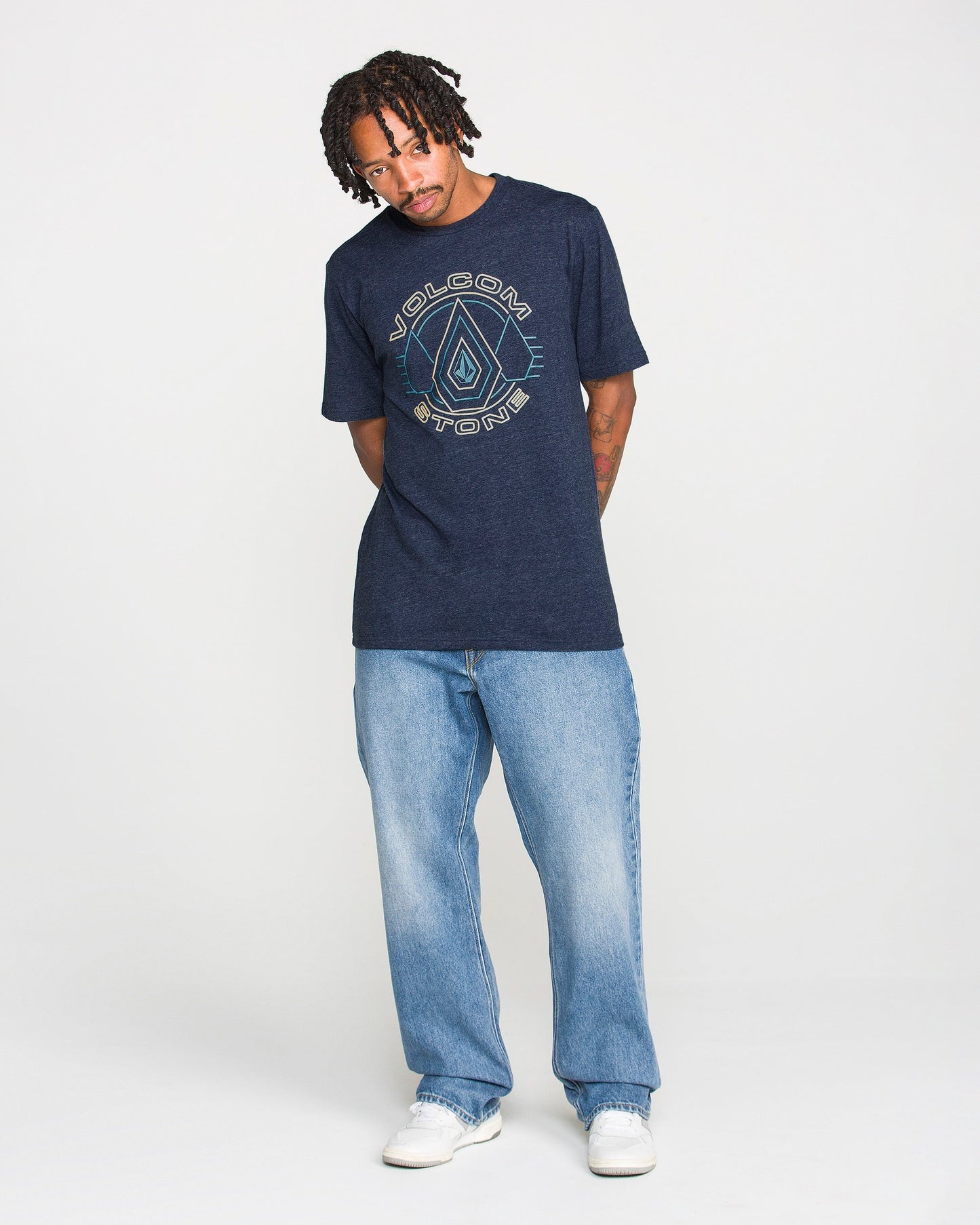 Man with short dreadlocks wears a navy Volcom Men's Line Feld Short Sleeve Tee, crafted from soft ring spun cotton, paired with light blue jeans and white sneakers, standing against a plain white background.