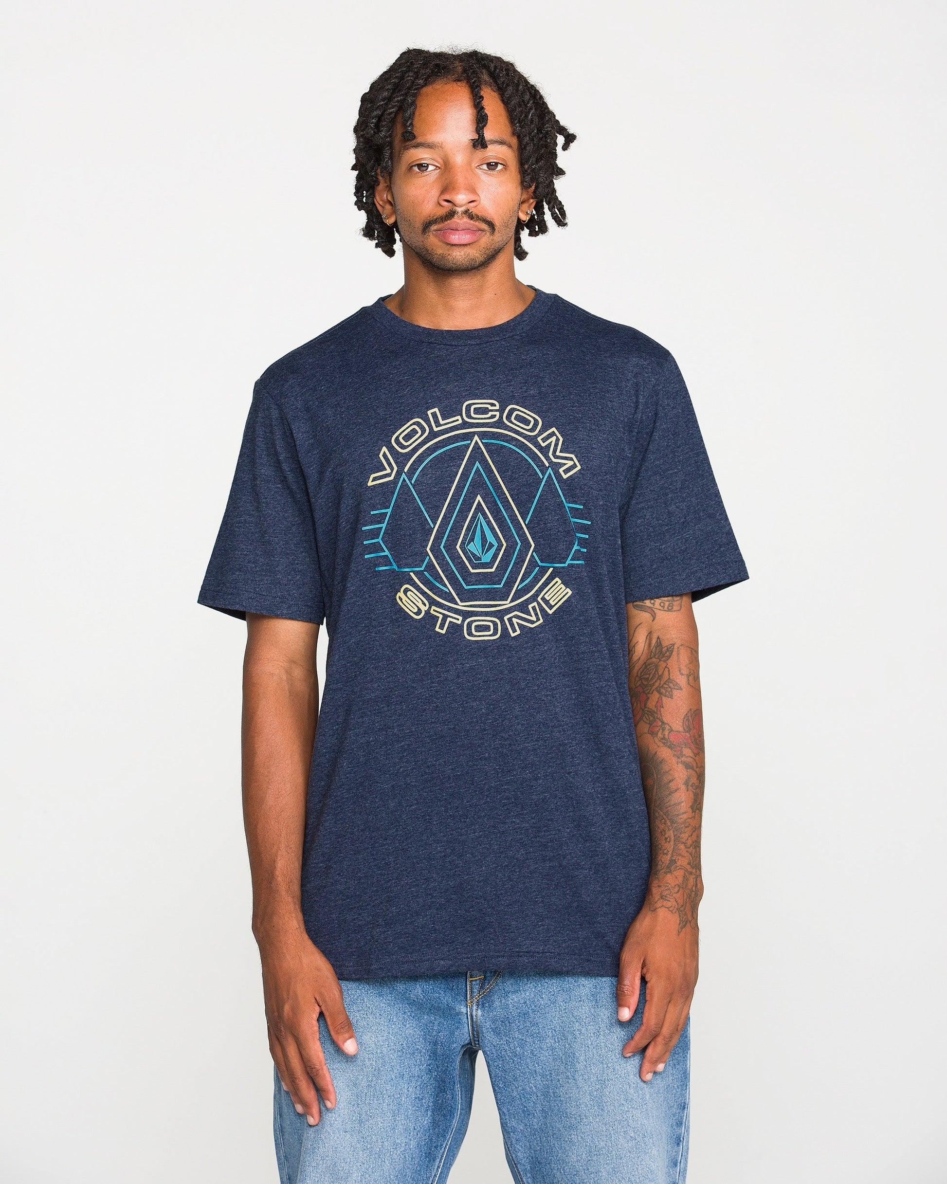 A person wearing a Volcom Men's Line Feld Short Sleeve Tee in navy blue heather, made from soft ring spun cotton, and light blue jeans stands against a plain white background, looking at the camera with a neutral expression.