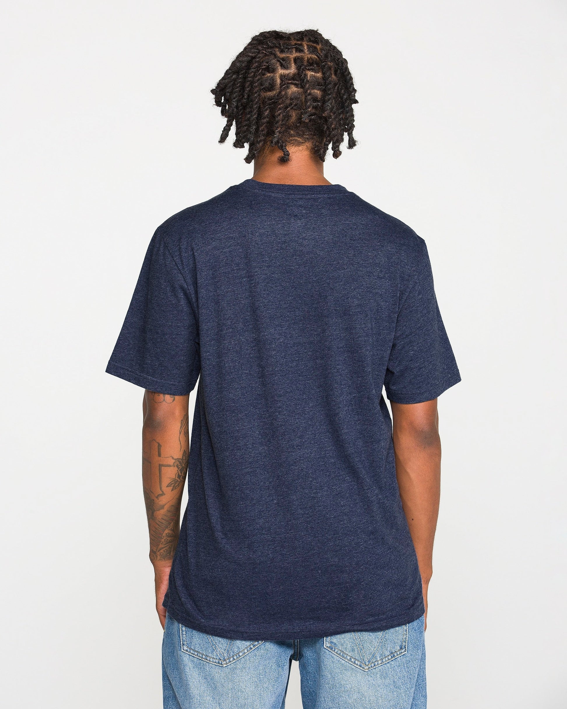 A person with short twists is standing facing away from the camera against a plain white background, wearing the Volcom Men's Line Feld Short Sleeve Tee in dark blue and light blue jeans.