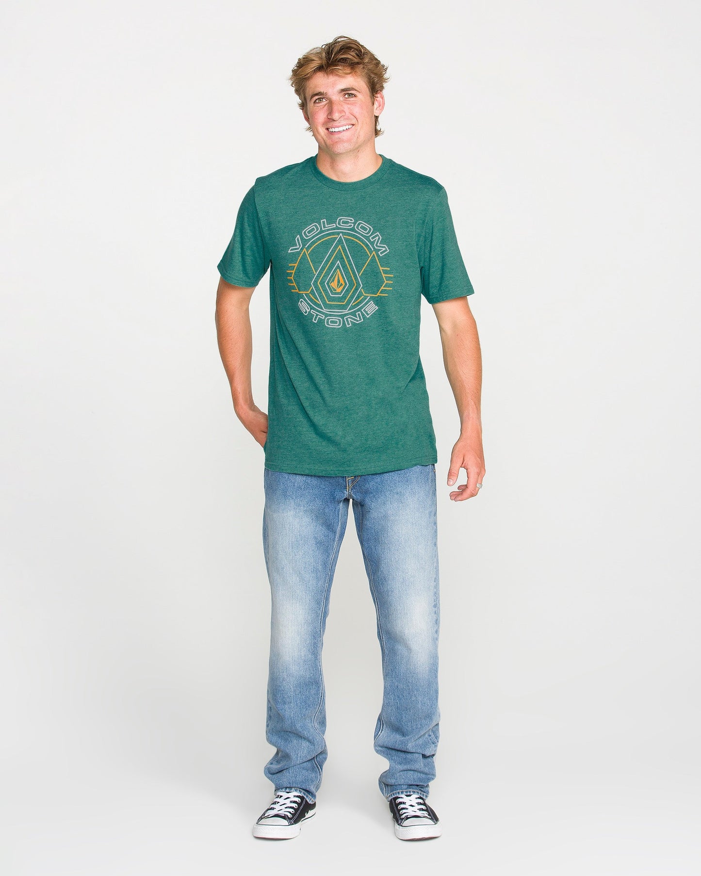 A smiling person stands facing forward, wearing the Volcom Men's Line Feld Short Sleeve Tee in Miami Green, featuring a soft-hand screen print, paired with light blue jeans and black sneakers, against a plain white background.