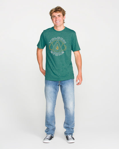 A smiling person stands facing forward, wearing the Volcom Men's Line Feld Short Sleeve Tee in Miami Green, featuring a soft-hand screen print, paired with light blue jeans and black sneakers, against a plain white background.