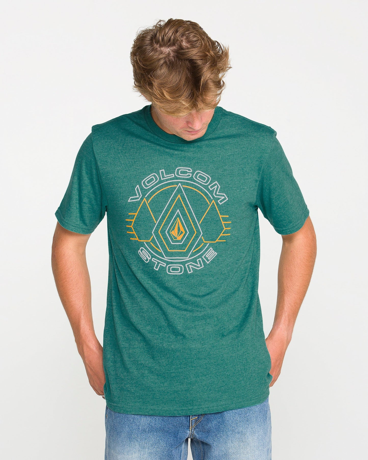 Wearing the Volcom Men's Line Feld Short Sleeve Tee in Miami Green, which features a Volcom Stone soft-hand screen print, and blue jeans, a person stands against a plain white background looking down with hands in their pockets.