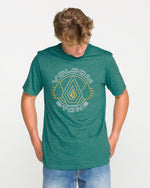Wearing the Volcom Men's Line Feld Short Sleeve Tee in Miami Green, which features a Volcom Stone soft-hand screen print, and blue jeans, a person stands against a plain white background looking down with hands in their pockets.
