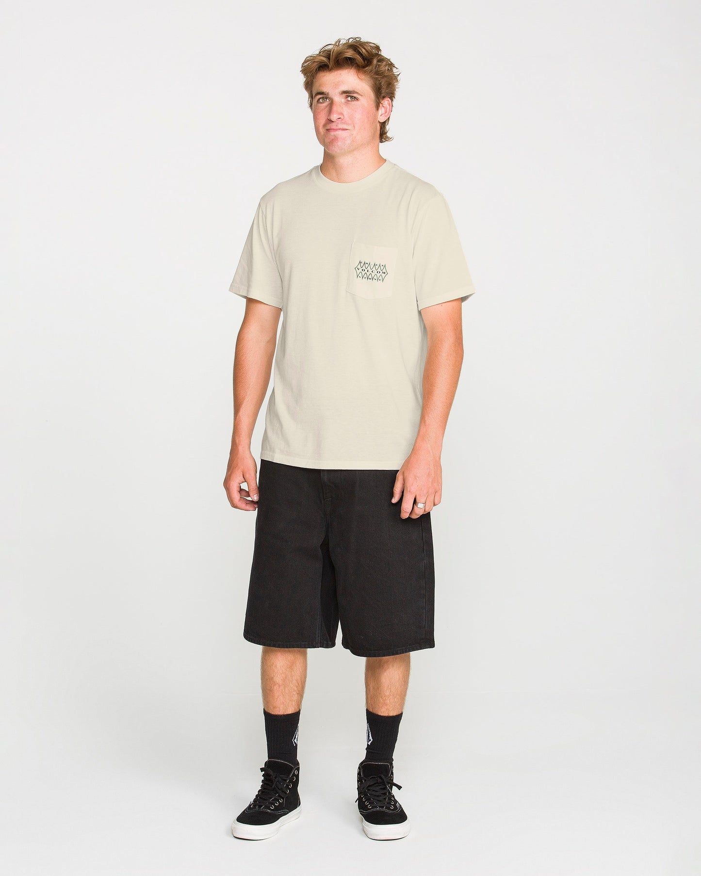 A person stands facing forward wearing the Volcom Men's Smokers Lounge Short Sleeve Tee in light beige, paired with black knee-length shorts, black crew socks, and black sneakers against a plain white background.