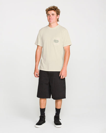 A person stands facing forward wearing the Volcom Men's Smokers Lounge Short Sleeve Tee in light beige, paired with black knee-length shorts, black crew socks, and black sneakers against a plain white background.