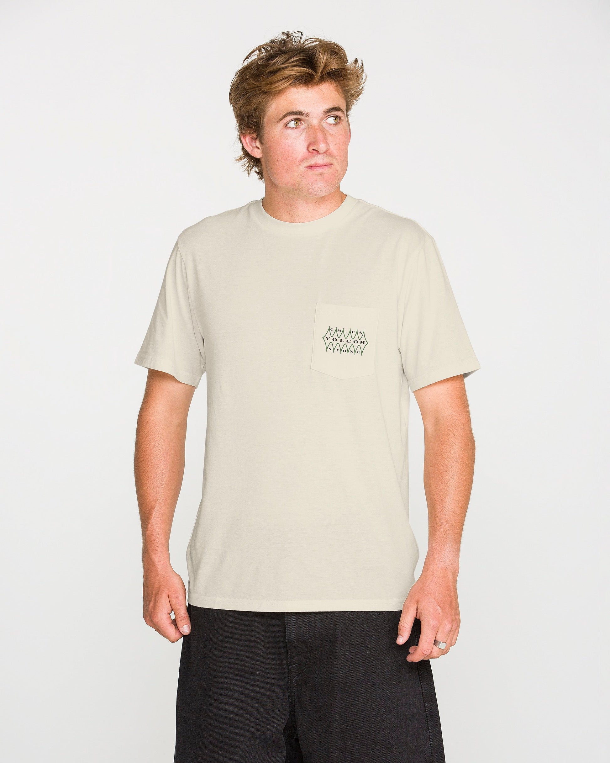 A person is wearing the Volcom Men's Smokers Lounge Short Sleeve Tee—a light beige crew neck t-shirt featuring a small geometric design on the pocket—paired with black shorts, standing against a plain white background and looking slightly to their right.