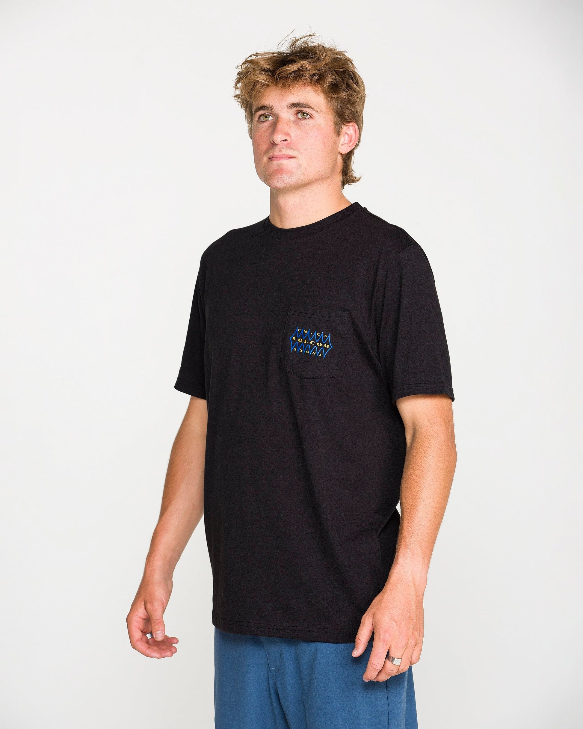 A young man with tousled hair wears the Volcom Men's Smokers Lounge Short Sleeve Tee in washed black heather, featuring a small chest pocket with a subtle pattern. He pairs it with blue pants and stands against a plain white background, looking slightly upward and to the side.