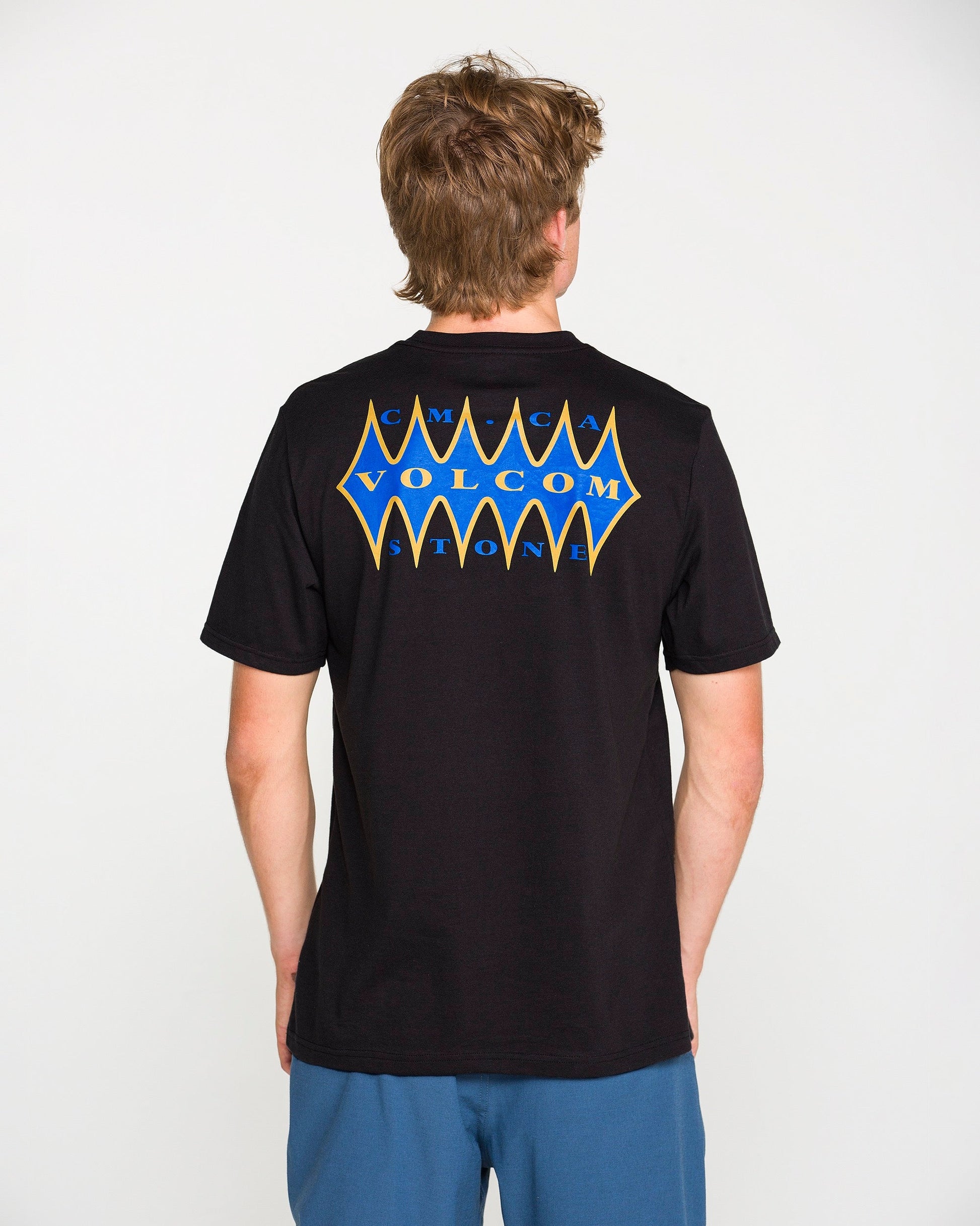 A person with short brown hair, wearing the Volcom Men's Smokers Lounge Short Sleeve Tee in washed black heather—with a blue and yellow Volcom Stone graphic on the back—stands facing away from the camera.