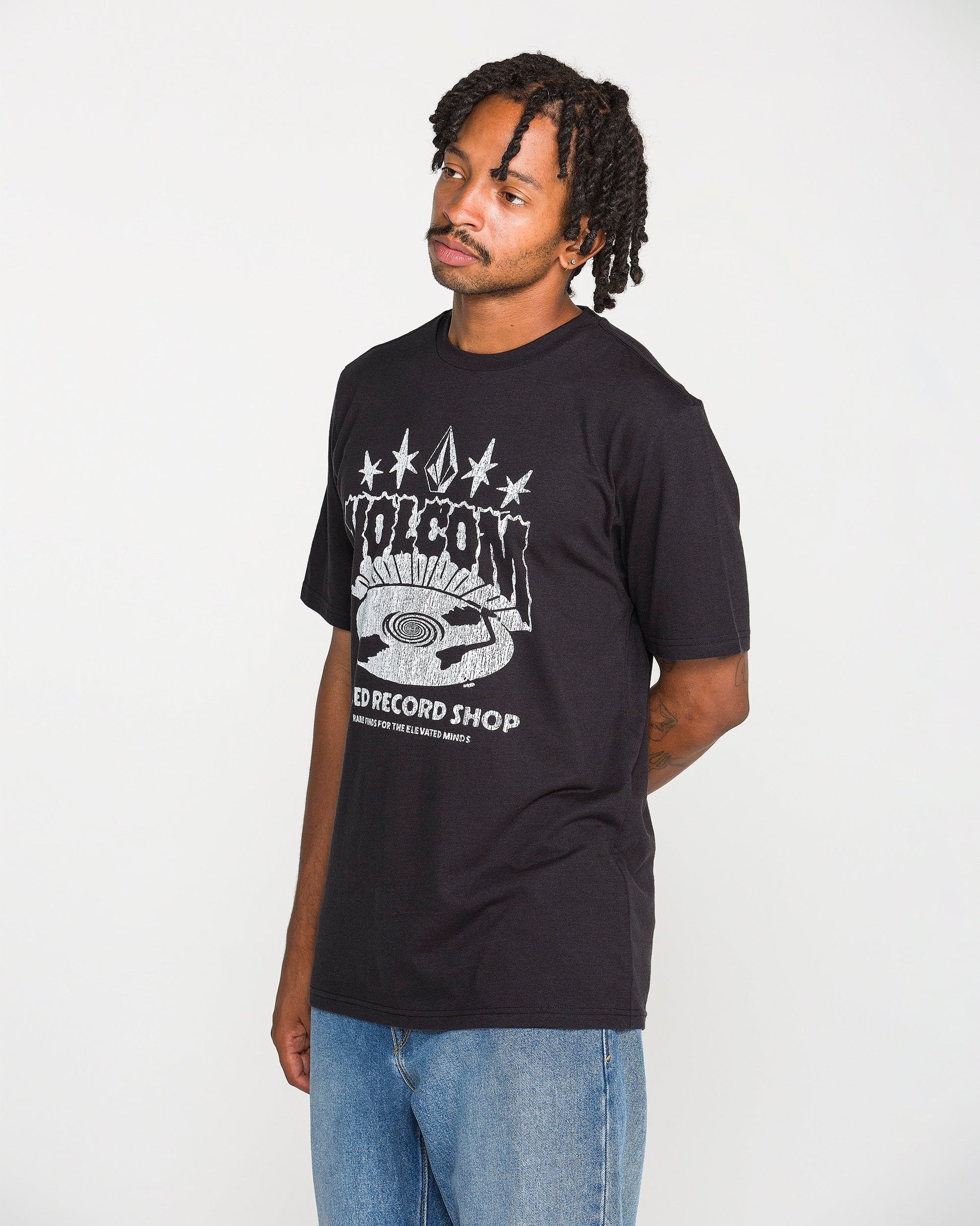 A man with short dreadlocks is wearing the Volcom Men's Collector Short Sleeve Tee in washed black, featuring crackle-ink "POISON RECORD SHOP" artwork, paired with blue jeans. He stands against a plain white background with his hands behind his back, looking slightly to the side.