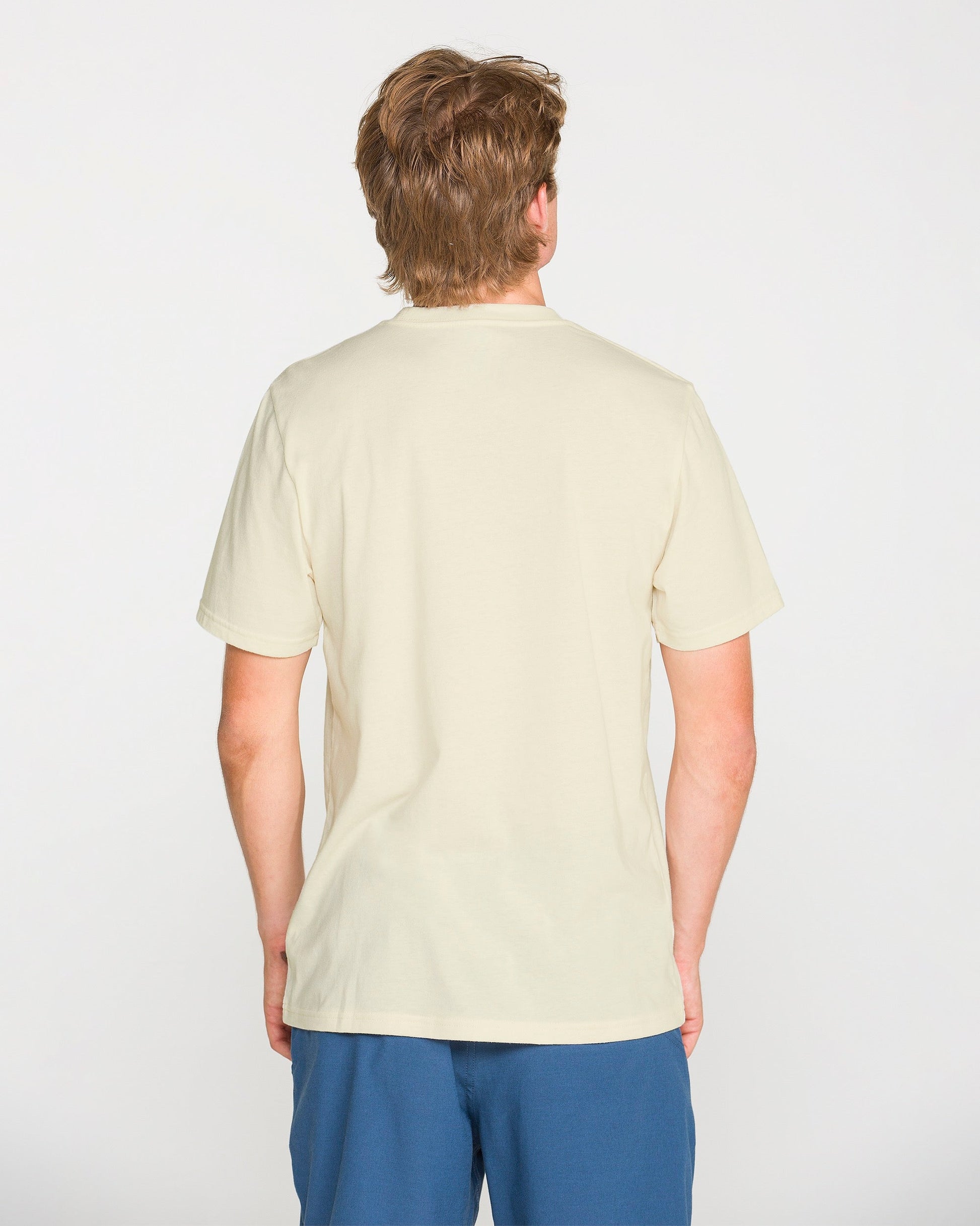 A person with light brown hair is shown from behind, wearing the Volcom Men's Filled Out Short Sleeve Tee in plain off-white and blue shorts, standing against a white background.