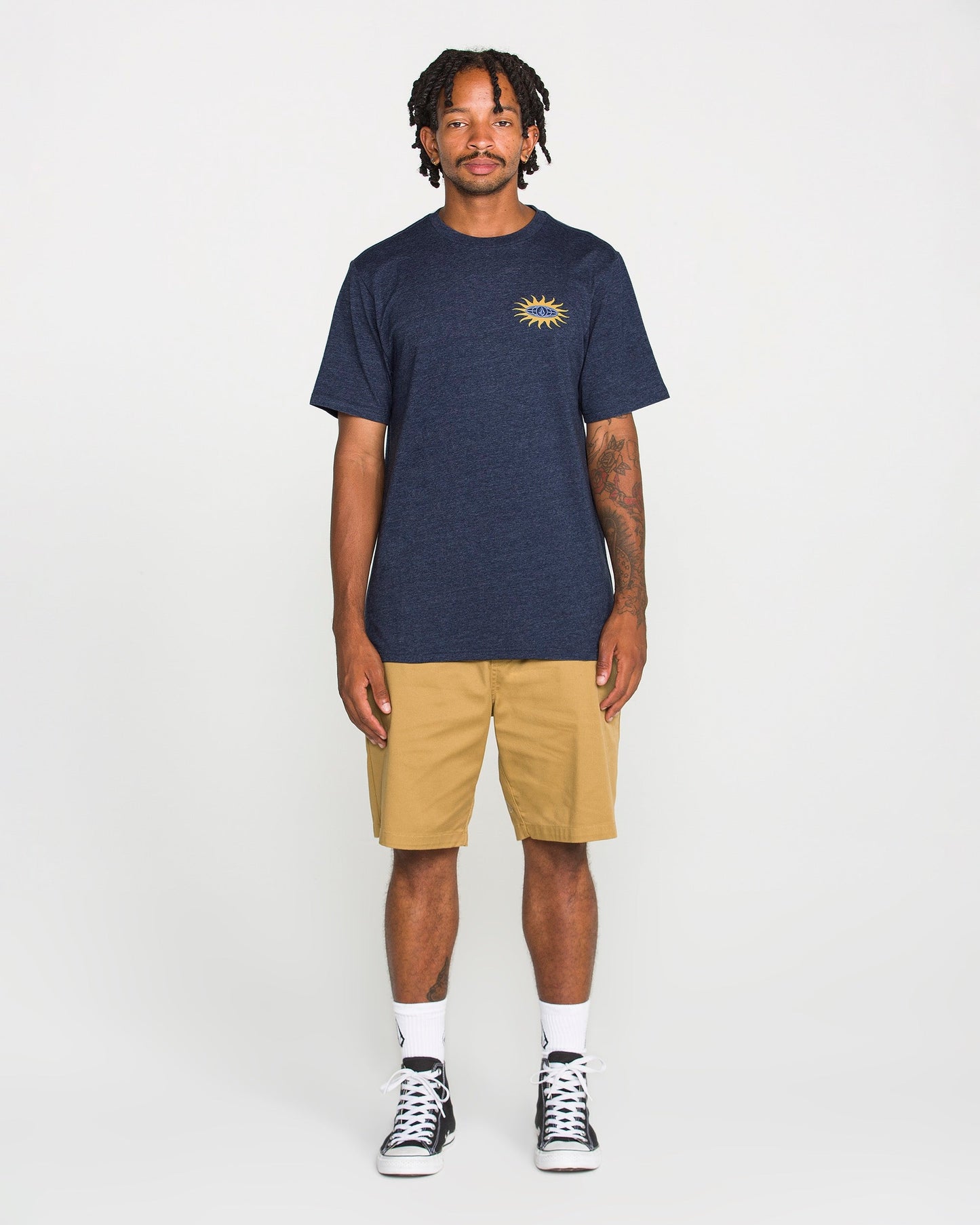 A person stands facing forward, wearing a Volcom Men's Spf91 Short Sleeve Tee in navy with a sun graphic on the chest, paired with khaki shorts, white socks, and black high-top sneakers. The background is plain white.