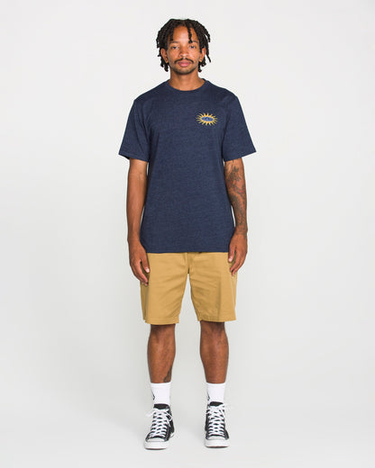 A person stands facing forward, wearing a Volcom Men's Spf91 Short Sleeve Tee in navy with a sun graphic on the chest, paired with khaki shorts, white socks, and black high-top sneakers. The background is plain white.
