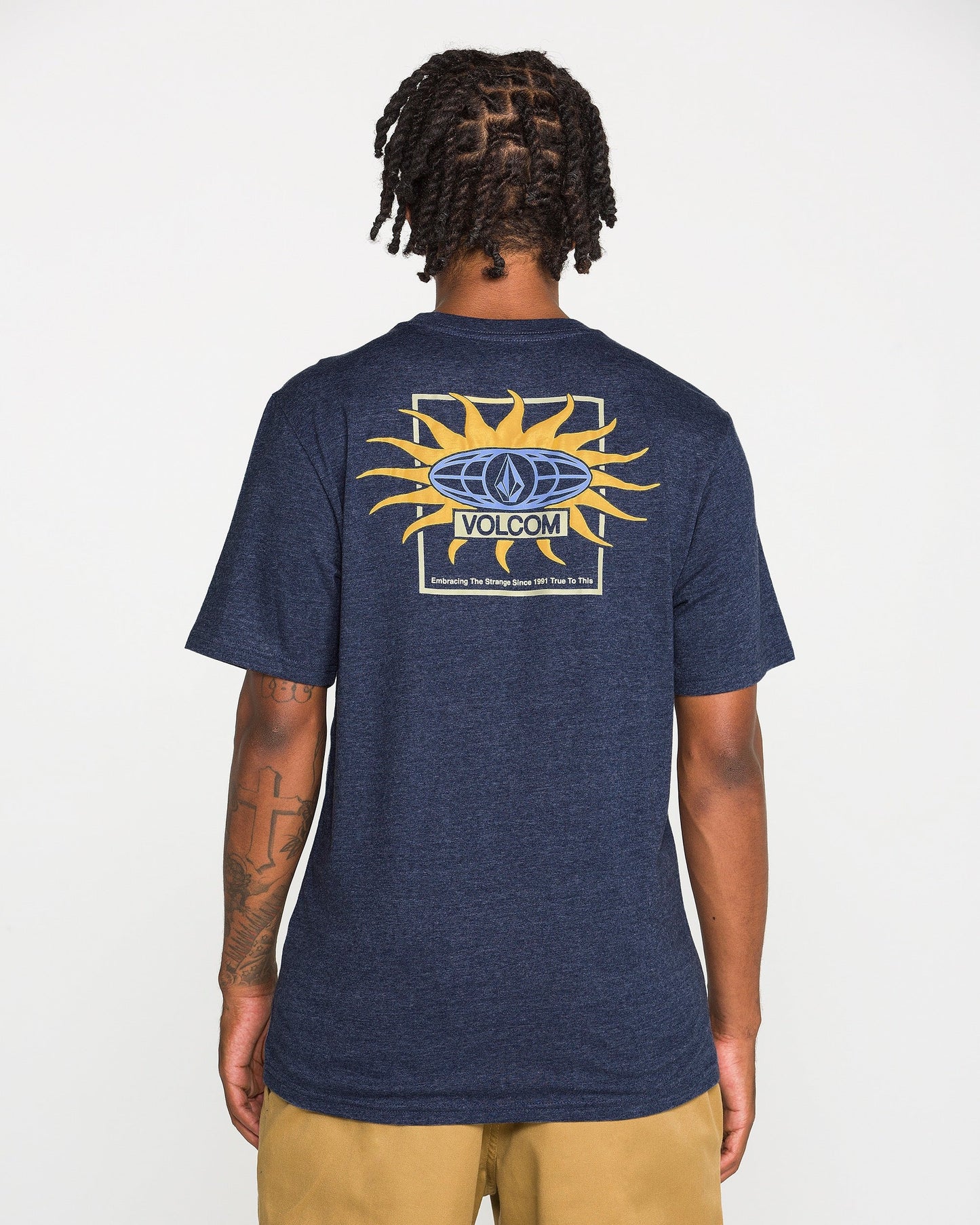 A person faces away, wearing the Volcom Men's Spf91 Short Sleeve Tee in navy blue, featuring a sun graphic and logo on the back. The medium-weight heather tee is paired with tan shorts, standing out against a plain white background.