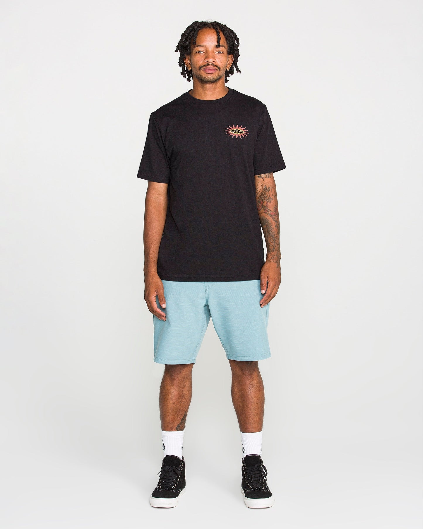 A man stands facing forward against a plain white background, wearing the Volcom Men's Spf91 Short Sleeve Tee—a medium-weight heather shirt with a small sun graphic—paired with light blue shorts, white socks, and black sneakers. The tee features a modern fit and is crafted from a soft ring spun cotton-poly blend for everyday comfort.