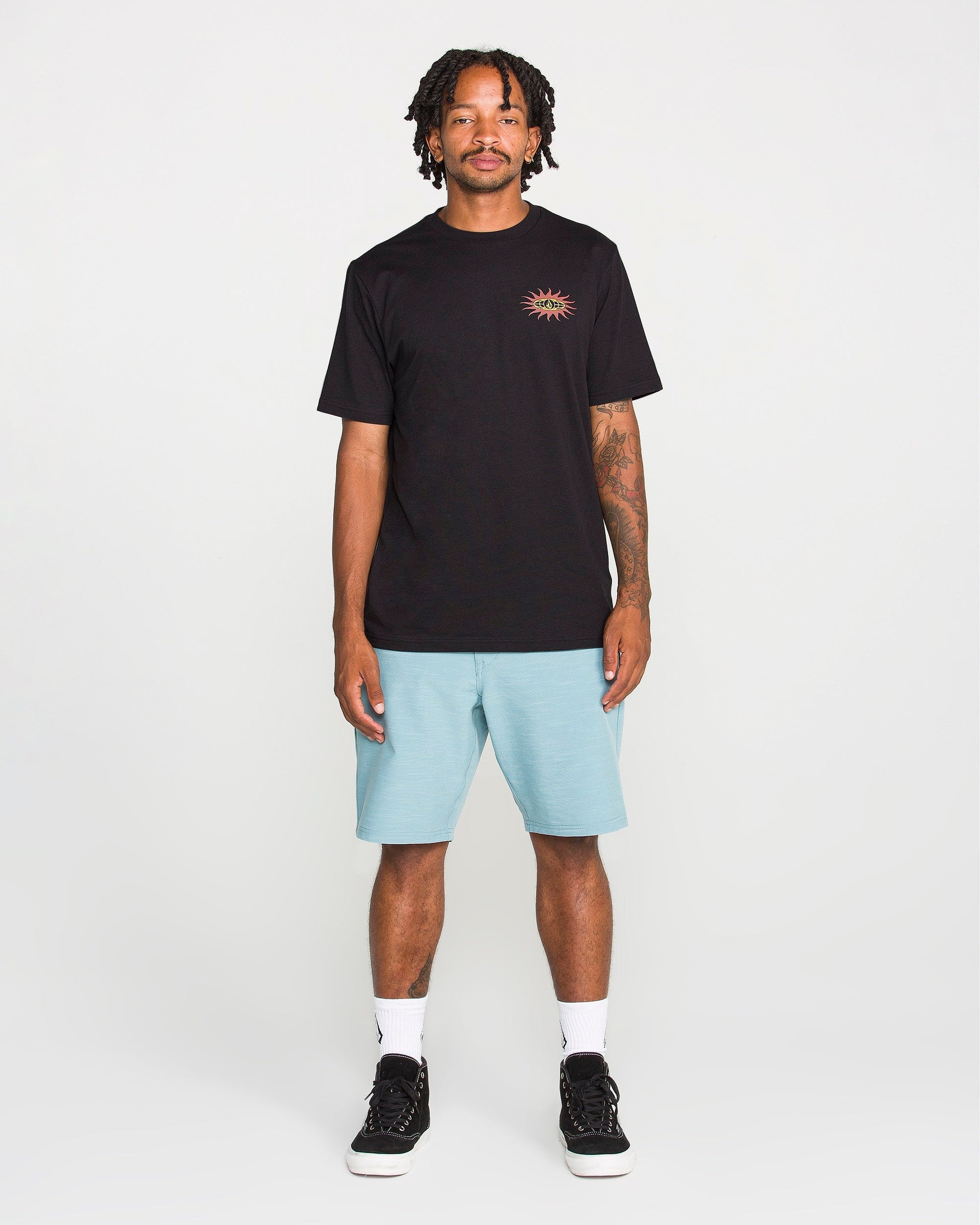 A man stands facing forward against a plain white background, wearing the Volcom Men's Spf91 Short Sleeve Tee—a medium-weight heather shirt with a small sun graphic—paired with light blue shorts, white socks, and black sneakers. The tee features a modern fit and is crafted from a soft ring spun cotton-poly blend for everyday comfort.