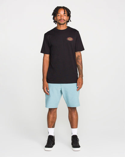 A man stands facing forward against a plain white background, wearing the Volcom Men's Spf91 Short Sleeve Tee—a medium-weight heather shirt with a small sun graphic—paired with light blue shorts, white socks, and black sneakers. The tee features a modern fit and is crafted from a soft ring spun cotton-poly blend for everyday comfort.