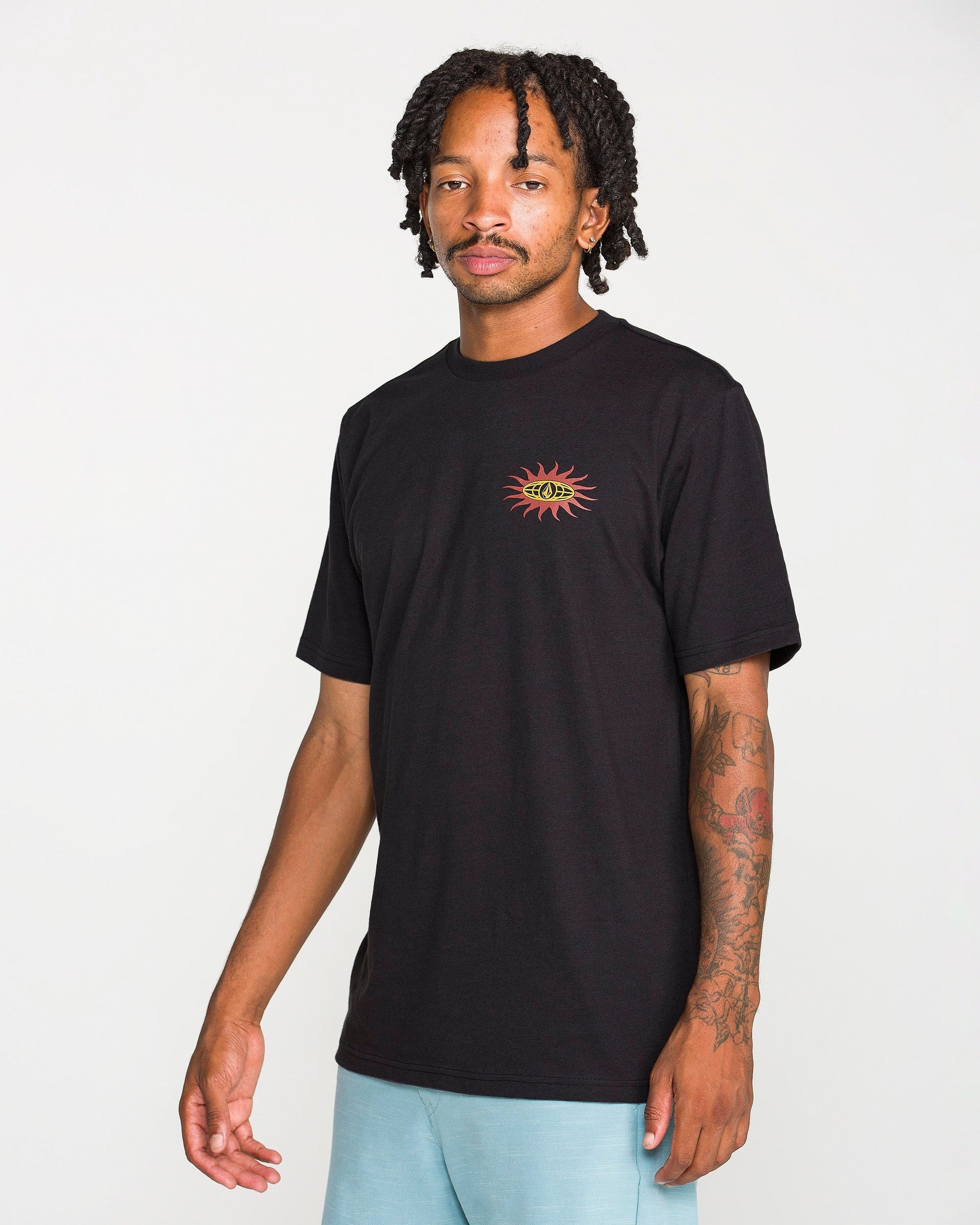 A man with short dreadlocks is wearing the Volcom Men's Spf91 Short Sleeve Tee in a black, medium-weight heather. The shirt features a small sun and eye graphic on the chest and is designed with a modern fit, crafted from a soft ring spun cotton-poly blend for everyday comfort. He stands against a plain white background.
