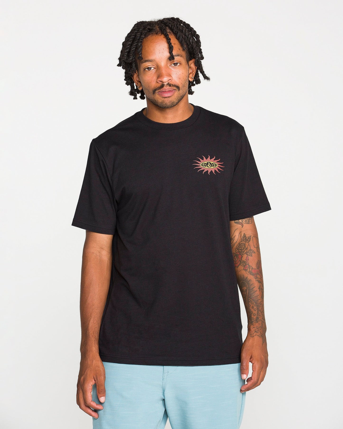 A person with short dreadlocks wears the Volcom Men's Spf91 Short Sleeve Tee, a black, medium-weight heather t-shirt with a small colorful sun graphic on the chest, paired with light blue shorts and standing against a plain white background. This modern fit tee is crafted from a soft ringspun cotton-poly blend, delivering both comfort and style.