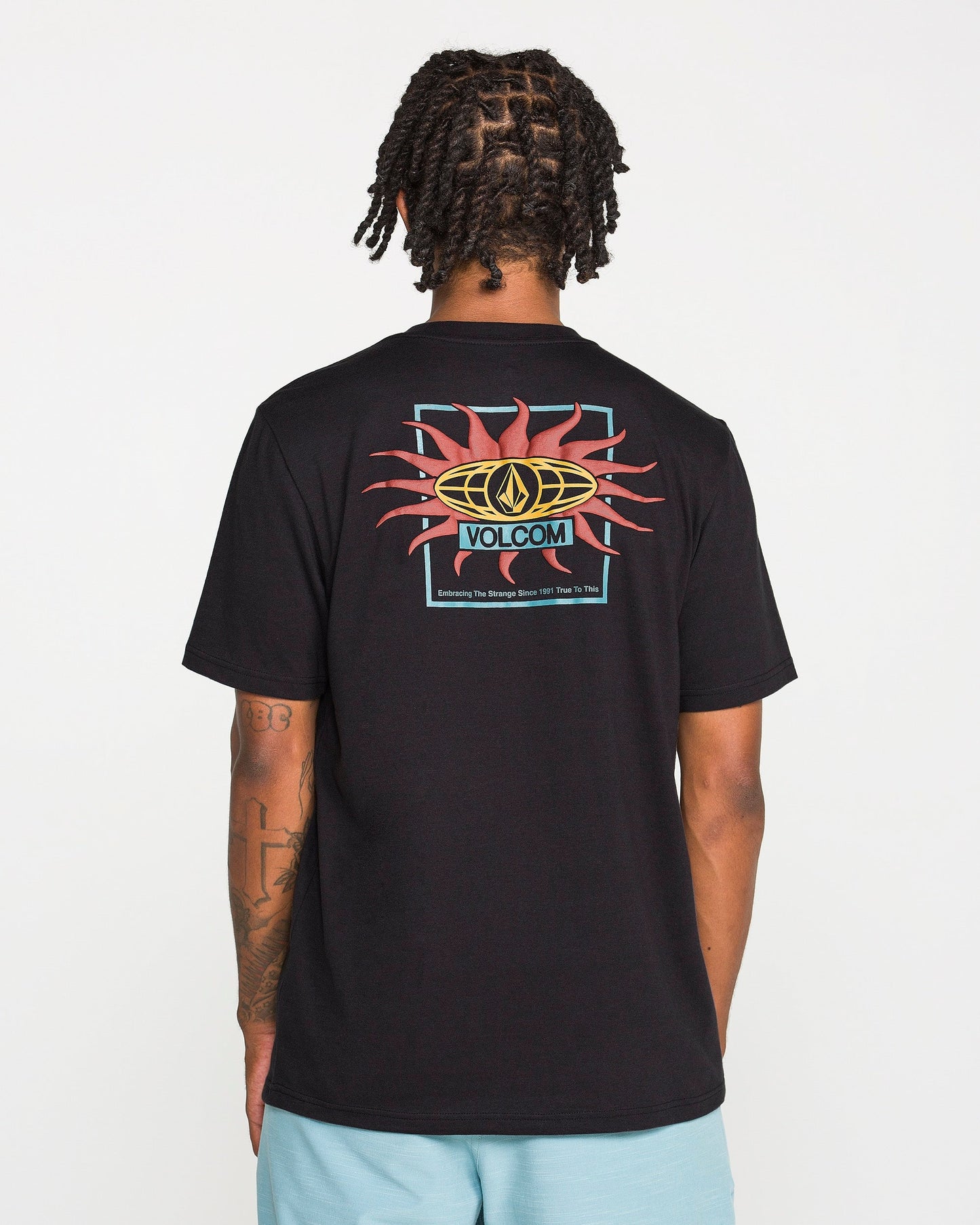 A person with braided hair is seen from behind wearing the Volcom Men's Spf91 Short Sleeve Tee in black heather, which features a vibrant back graphic showcasing the word "Volcom" and a sun motif. Crafted from a soft ring spun cotton-poly blend in a modern fit, this medium-weight T-shirt pairs seamlessly with light blue shorts.