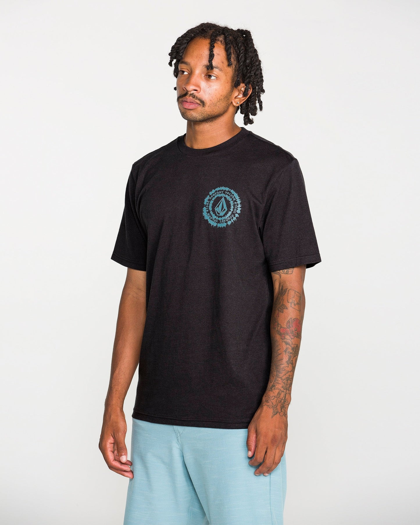 A man with short dreadlocks wears the Volcom Men's Still Spiraling Short Sleeve Tee in washed black, which features a small blue graphic done in a soft hand screen print on the left chest. He pairs it with light blue shorts and stands against a plain white background.