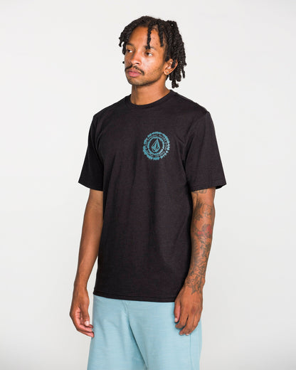 A man with short dreadlocks wears the Volcom Men's Still Spiraling Short Sleeve Tee in washed black, which features a small blue graphic done in a soft hand screen print on the left chest. He pairs it with light blue shorts and stands against a plain white background.