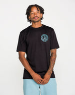 A tattooed man wears the Volcom Men's Still Spiraling Short Sleeve Tee in washed black, which features a blue circular logo on the chest, paired with light blue shorts. The soft hand screen print on the modern-fit heather tee gives a subtle, contemporary touch. He stands against a plain white background, looking at the camera with his hands clasped in front of him.