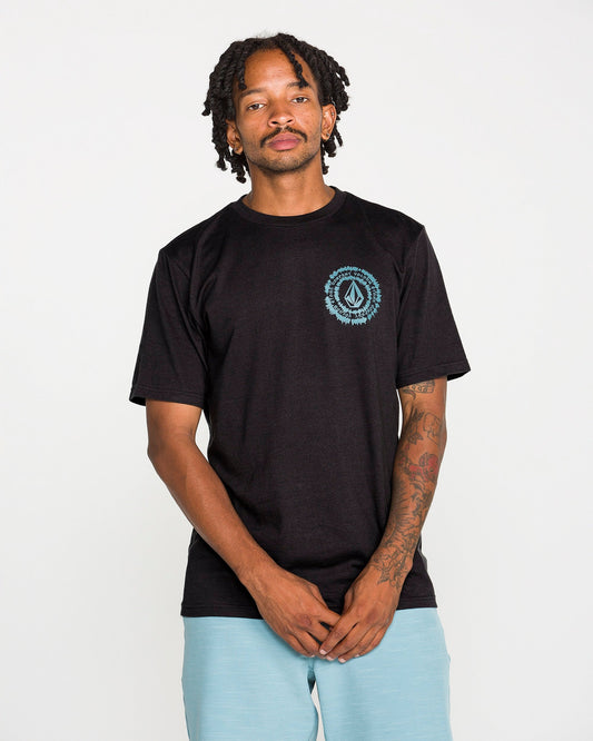 A tattooed man wears the Volcom Men's Still Spiraling Short Sleeve Tee in washed black, which features a blue circular logo on the chest, paired with light blue shorts. The soft hand screen print on the modern-fit heather tee gives a subtle, contemporary touch. He stands against a plain white background, looking at the camera with his hands clasped in front of him.